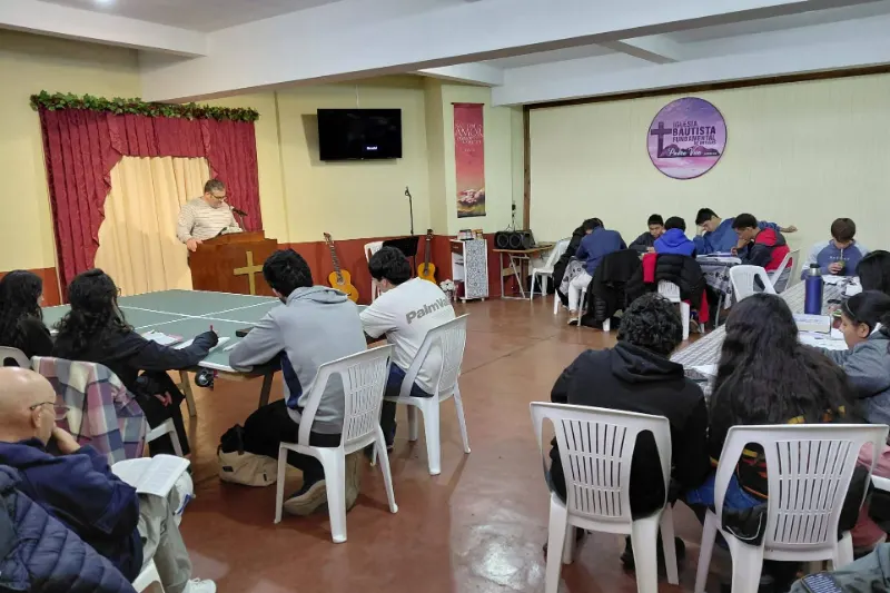 Jóvenes sentados alrededor de mesas con sus Biblias mientras el pastor enseña.