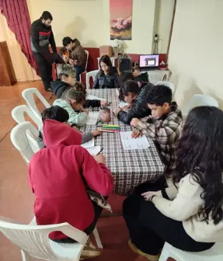 Niños pintando en la mesa mientras un joven toca la guitarra.