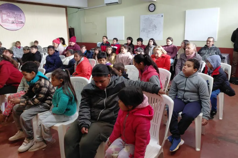 Niños y adultos sentados en sillas blancas durante una reunión en el salón de la iglesia.